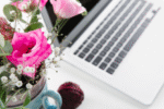 Close-up of a silver laptop on a white surface beside a ceramic mug holding a bouquet of bright pink and light pink flowers with greenery and baby’s breath. The setup creates a clean and feminine workspace aesthetic.