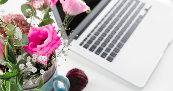 Close-up of a silver laptop on a white surface beside a ceramic mug holding a bouquet of bright pink and light pink flowers with greenery and baby’s breath. The setup creates a clean and feminine workspace aesthetic.