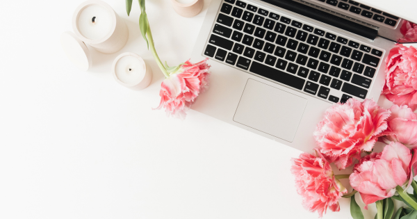 Flat lay of a laptop on a white desk surrounded by pink tulips and white candles. The flowers and candles create a soft, feminine workspace aesthetic.